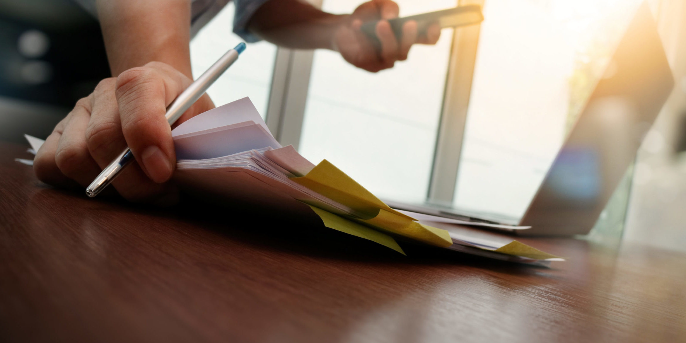 A desk with paperwork. A person's hards are visible. The hands are holding a pencil and phone while placing down more paperwork on the desk.