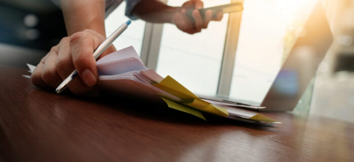 A desk with paperwork. A person's hards are visible. The hands are holding a pencil and phone while placing down more paperwork on the desk.