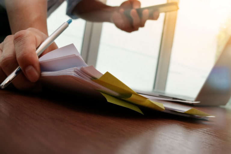 A desk with paperwork. A person's hards are visible. The hands are holding a pencil and phone while placing down more paperwork on the desk.