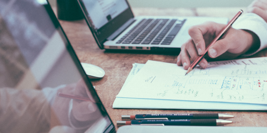 Untitled Design (3) Two laptops sit beside each other while hands come into the frame holding a pencil.