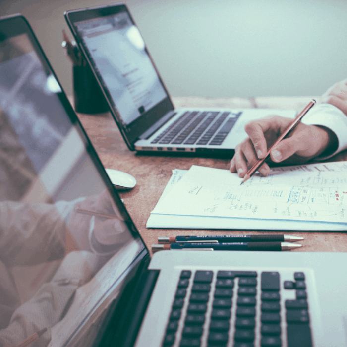 Two laptops sit beside each other while hands come into the frame holding a pencil.