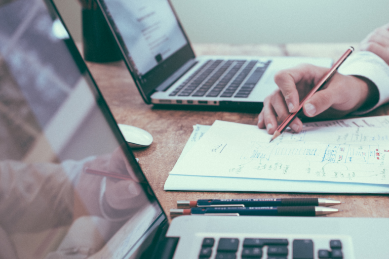Two laptops sit beside each other while hands come into the frame holding a pencil.