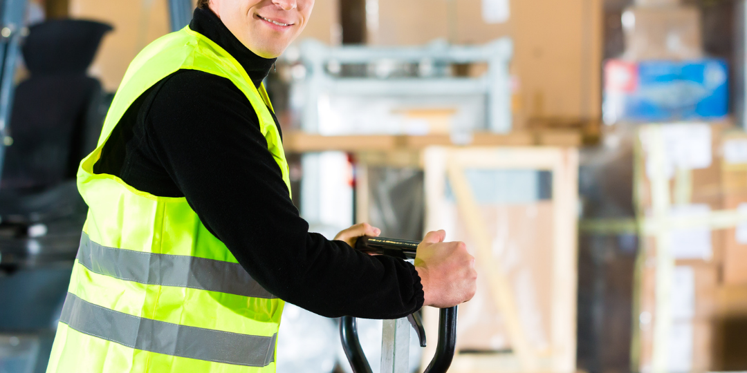 Untitled Design (4) Merchandise handler smiles at camera in front of stacks of boxes. A forklift is beside him