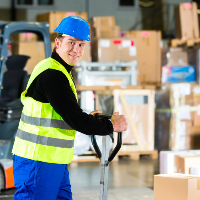 Untitled Design (4) Merchandise handler smiles at camera in front of stacks of boxes. A forklift is beside him