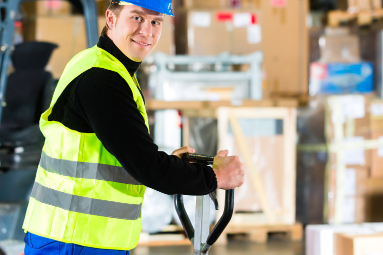 Merchandise handler smiles at camera in front of stacks of boxes. A forklift is beside him