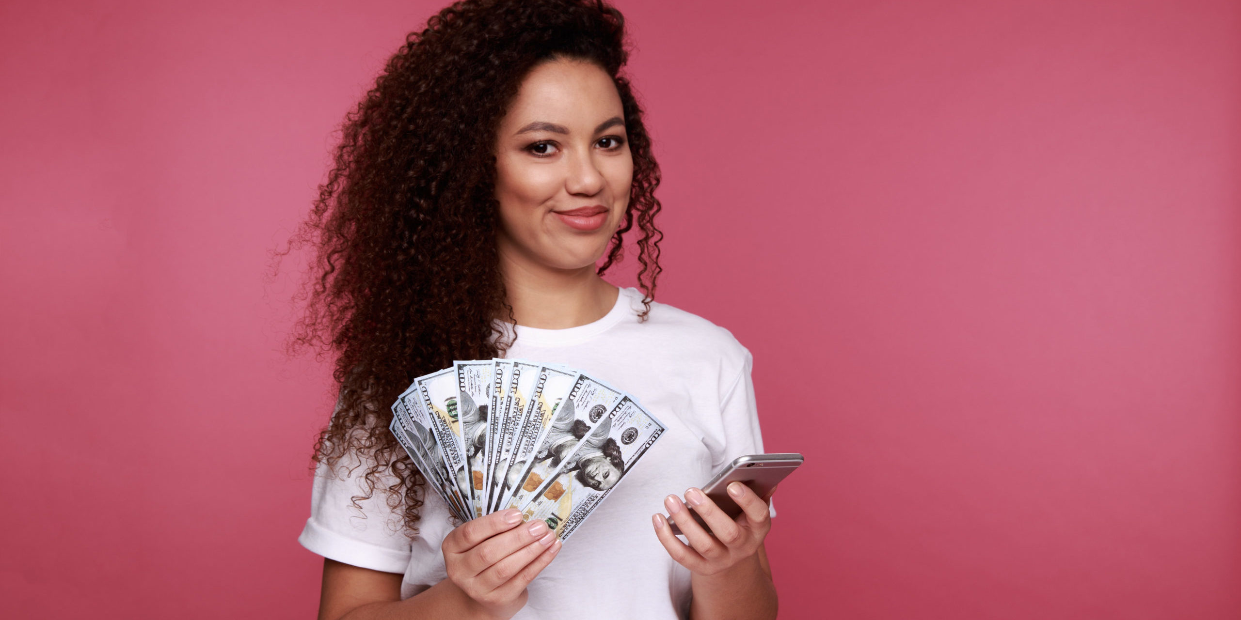 Portrait Of An Excited Young African Woman Holding Bunch Of Money Banknotes And Looking At Mobile Phone Isolated Over Pink Background