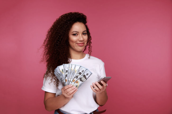 Portrait Of An Excited Young African Woman Holding Bunch Of Money Banknotes And Looking At Mobile Phone Isolated Over Pink Background