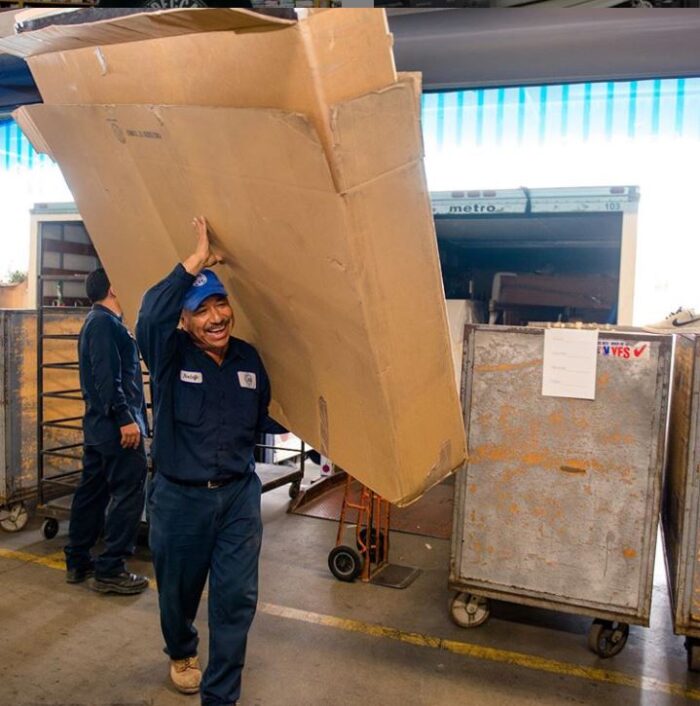 IMG-3496 Man smiles as he holds up a large box. Behind him is a large truck with many other large boxes.