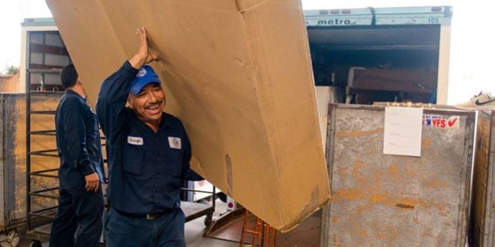 IMG-3496 Man smiles as he holds up a large box. Behind him is a large truck with many other large boxes.
