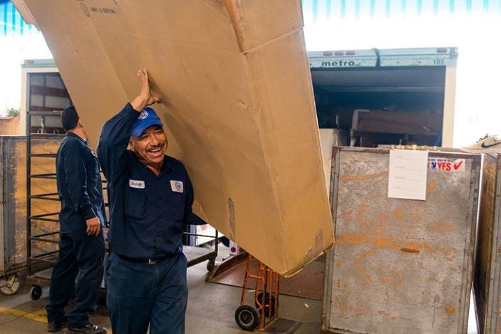 Man smiles as he holds up a large box. Behind him is a large truck with many other large boxes.
