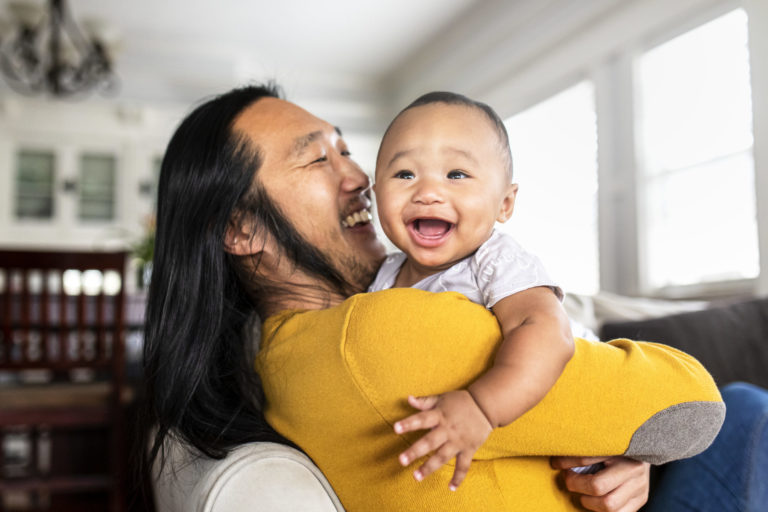 A smiling Asian father with long hair holds his laughing baby.