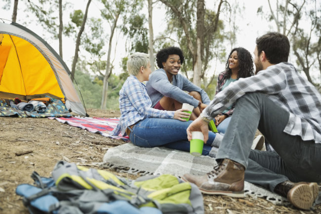 Friends talking while camping outdoors Friends sit on blankets on the ground outdoors. There are trees in the background and a yellow camping tent beside the friends.