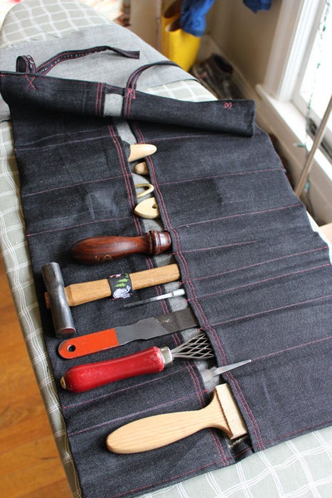 A denim roll case is rolled out onto an ironing table. The roll is filled with various tools like a paintbrush and hammer.