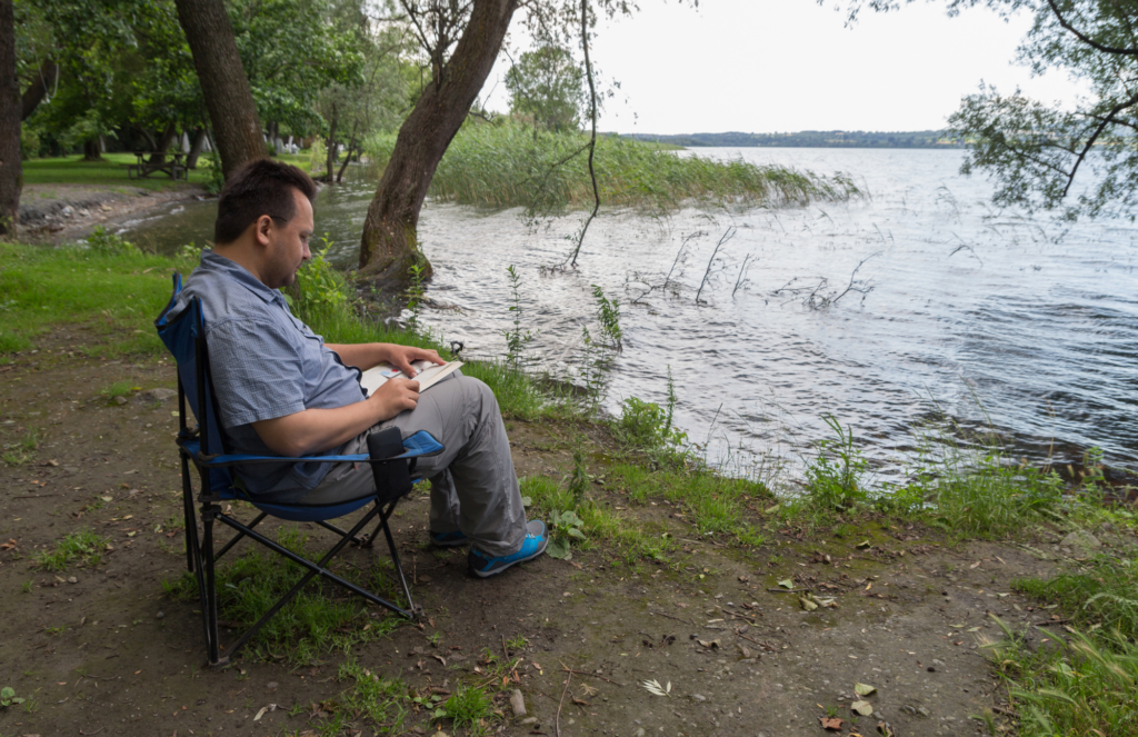 Man sits on a foldable cloth chair on the edge of a lake in the forest. He is reading a book on his lap.