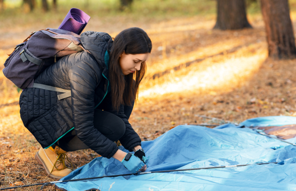 Woman packs up her blue camping tent. She is wearing a purple backpack and a black down jacket.